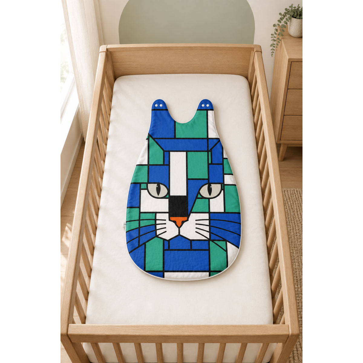 A baby sleep sack or sleeping bag with a colorful geometric cat face design, displayed on a white mattress inside a light wood crib.