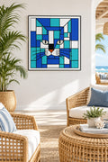 A framed square wall art print featuring a skismic cat face in shades of blue, teal, white, and black, hanging on a white wall in a bright coastal living room with wicker furniture and a plant.